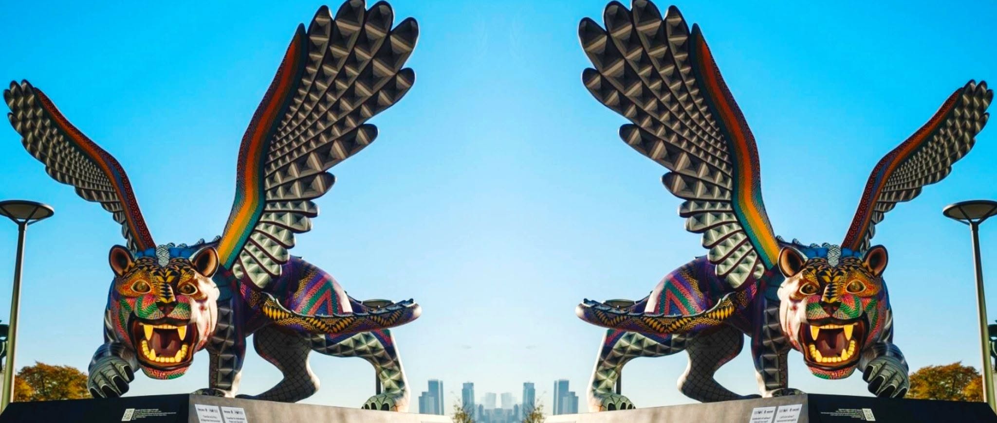Two mirrored, colorful winged jaguar sculptures against a blue sky.